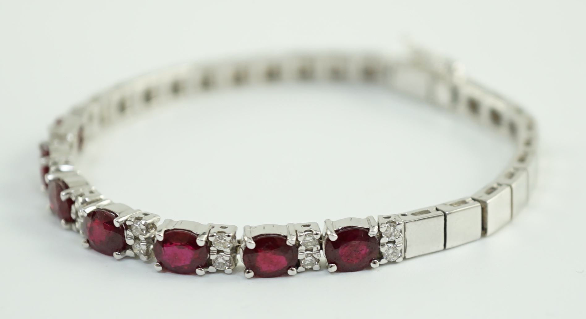 A 750 white gold and seven stone oval cut ruby set line bracelet, set with sixteen stone diamond spacers